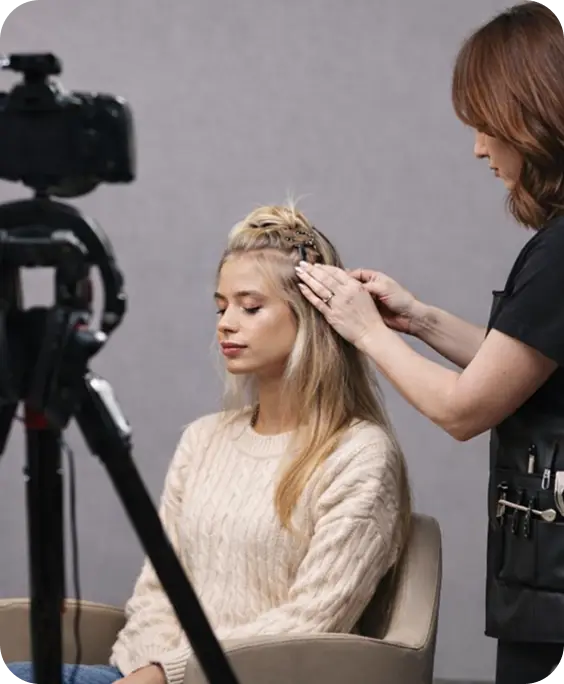 Blonde Frau wird vor einer Kamera von einer Stylistin frisiert