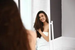 Beautiful young woman with healthy long hair looking in mirror at home